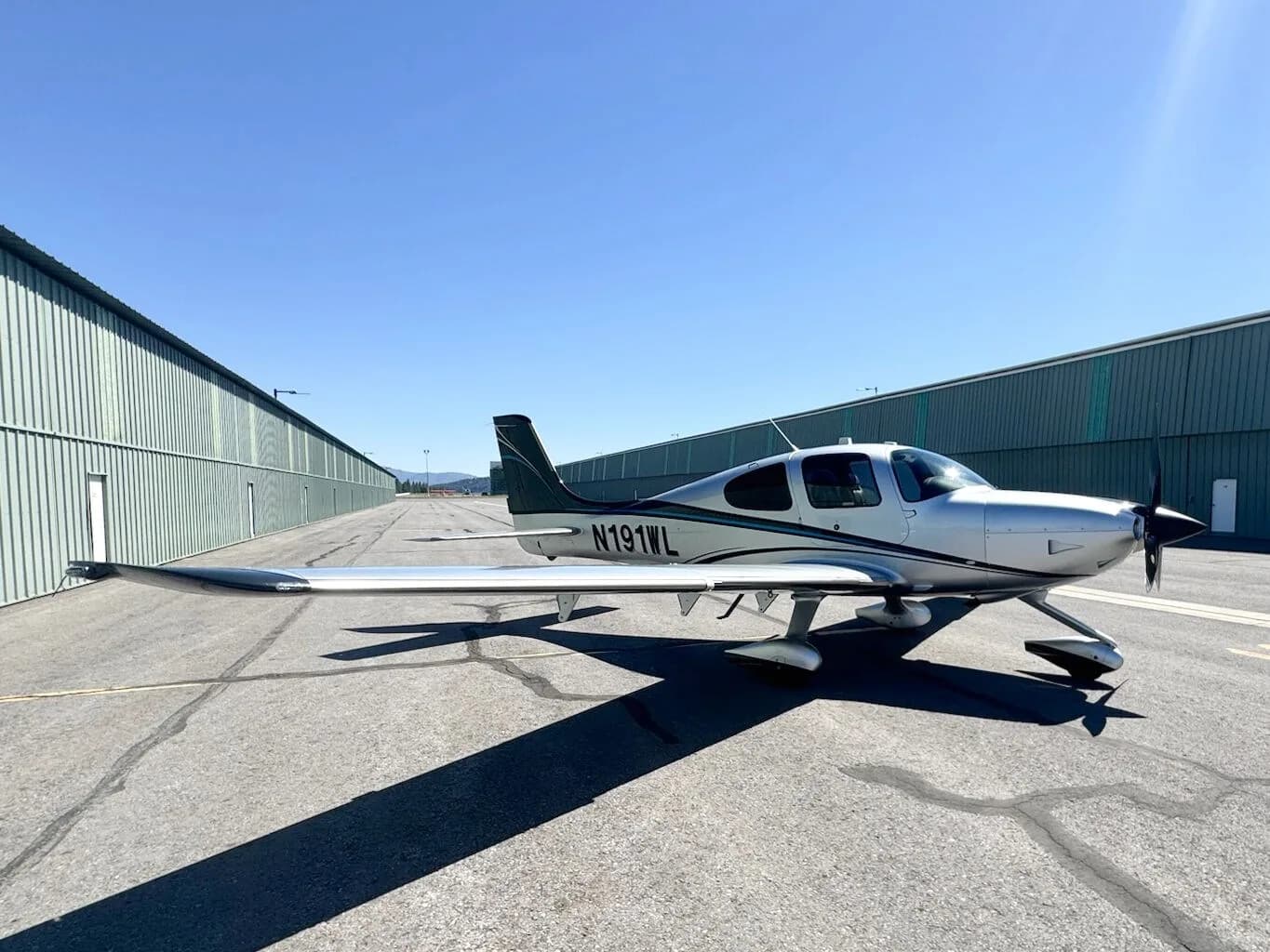A side view of a silver and black Cirrus SR22T aircraft with N191WL, parked on an asphalt apron next to hangars.
