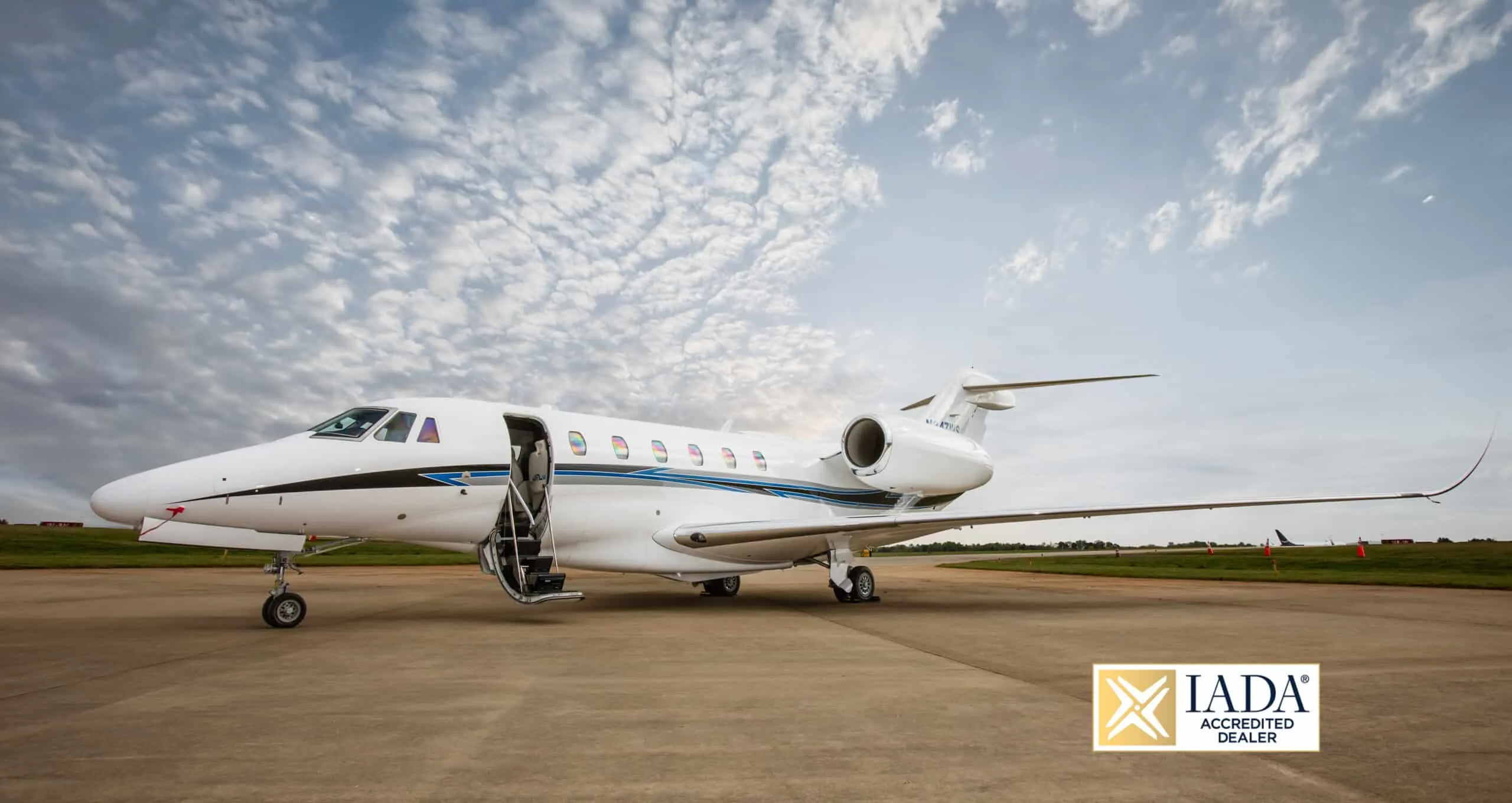 A white business jet with blue and black stripes on the fuselage, parked on a tarmac with its main door open.