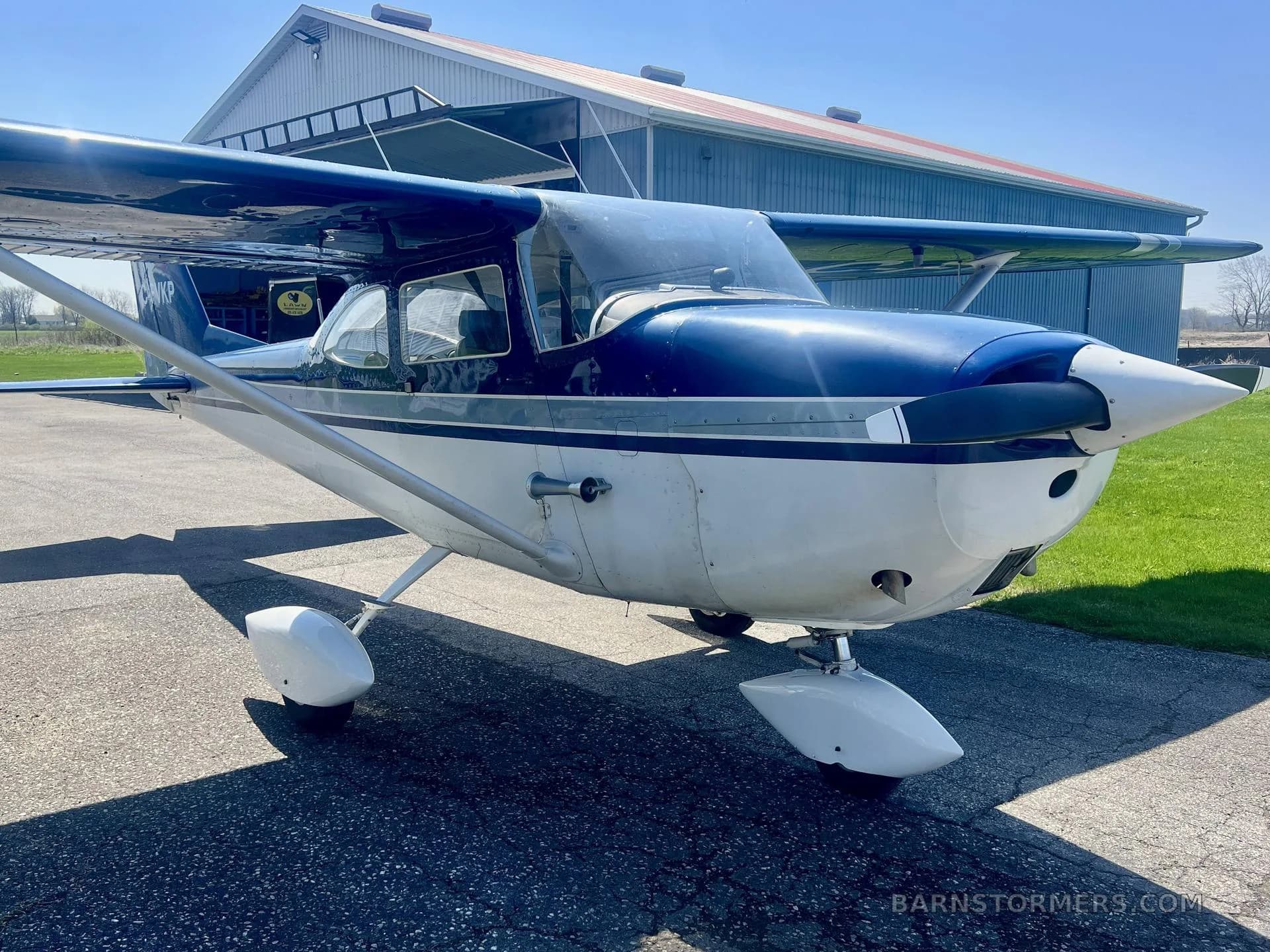 A blue and white 1967 Cessna 172H, registration C-FVKP, parked on an tarmac in front of a hangar, viewed from the front-left.