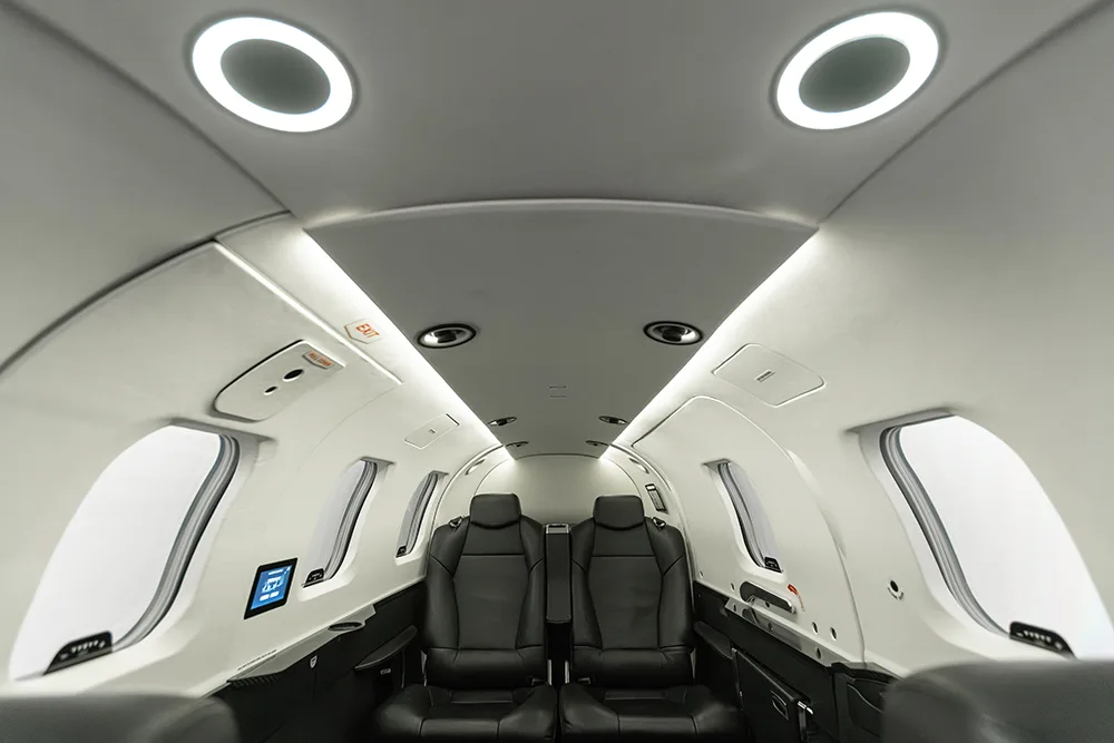 A bright private jet cabin interior with circular ceiling lights and black leather seats.