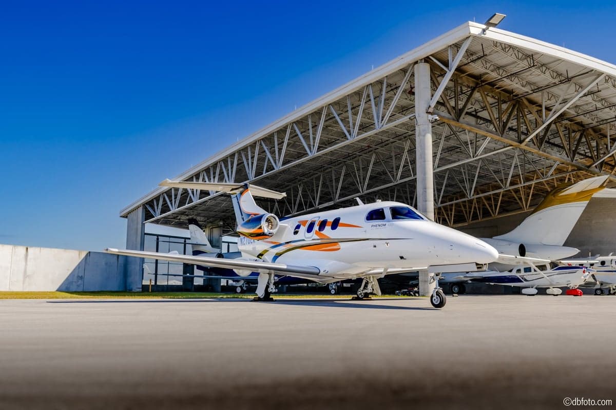 A Phenom 100 aircraft with a white, orange, and black paint scheme parked in front of a hangar.