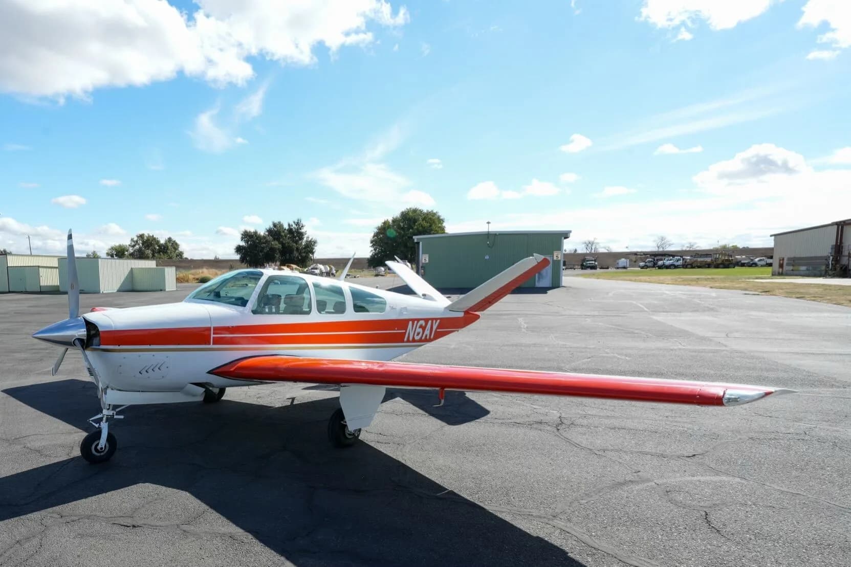 A low-angle view of the white and orange Beechcraft Bonanza N6AY on the tarmac, showcasing its full wing.