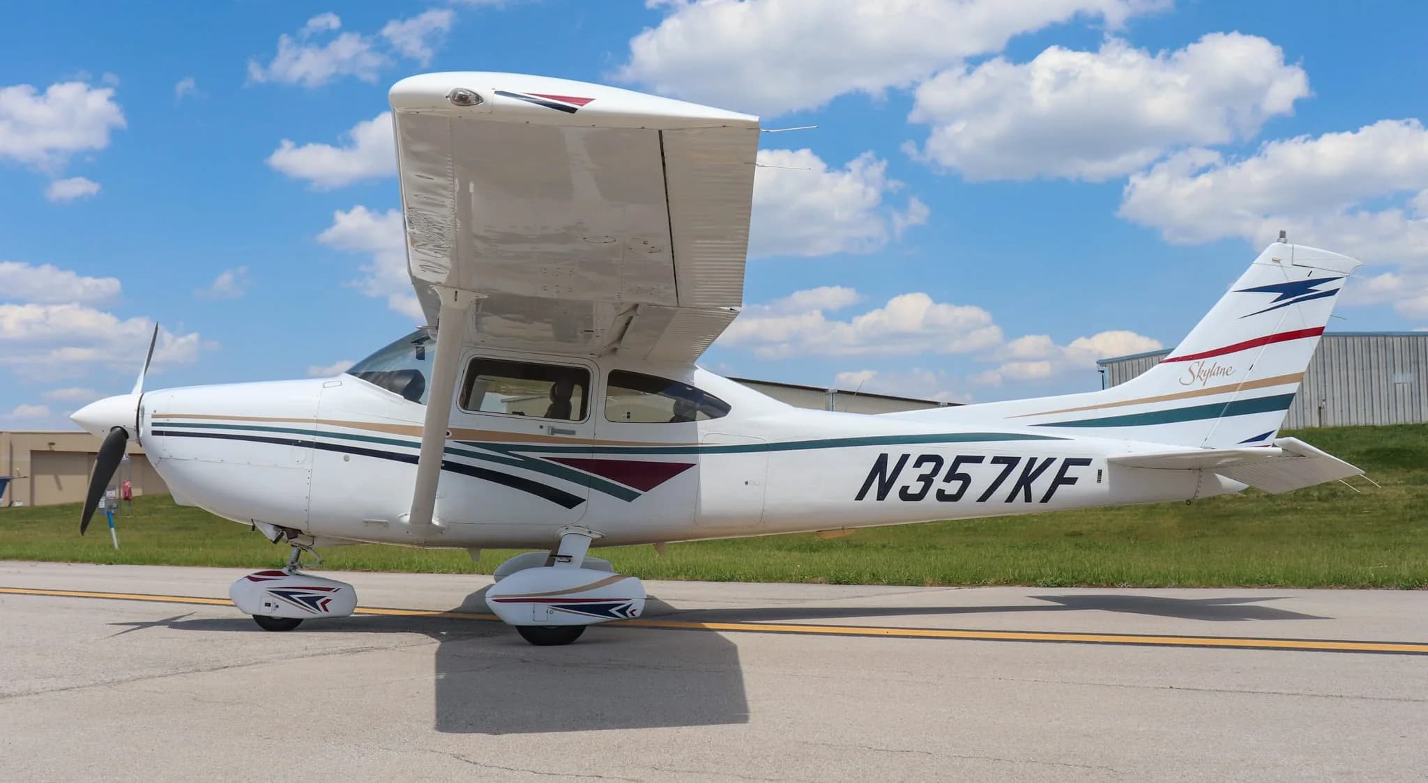 A side view of a white Cessna 182 Skylane with gold, green, and maroon stripes parked on an airport ramp.