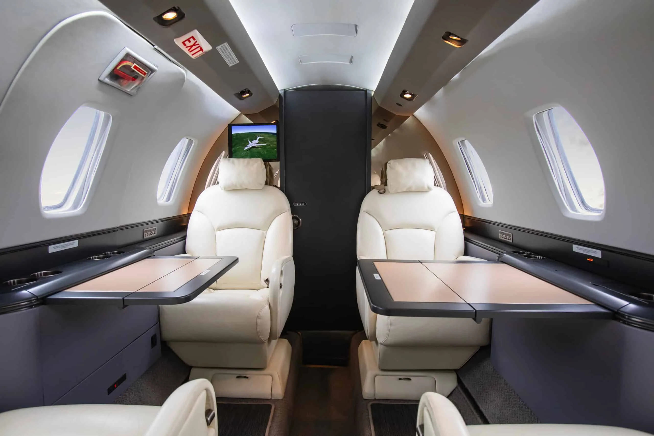 The interior of a private jet cabin showing light-colored leather seats, fold-out tables, and a screen at the rear.