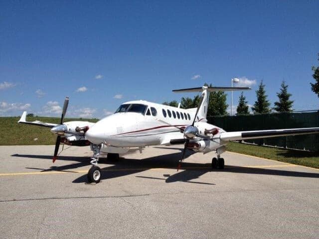 A white Beechcraft King Air with red and black stripes parked on an airport tarmac under a clear blue sky.