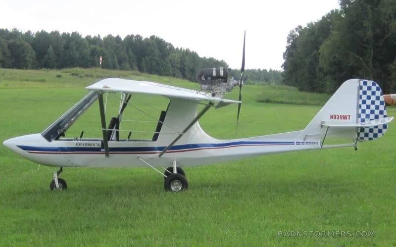 A white ultralight aircraft with blue and red stripes and a checkered tail, parked on a grassy field.