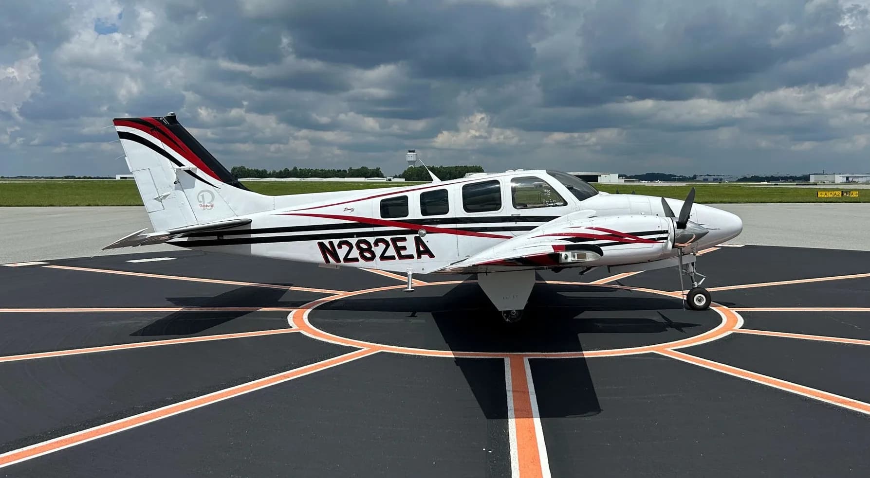 A white Beechcraft Bonanza N282EA with black and red stripes parked on an airport tarmac under a cloudy sky.
