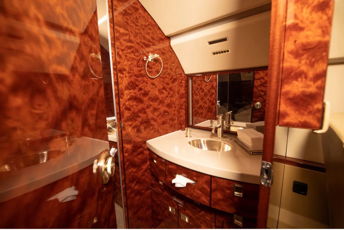 The aircraft's lavatory with a modern sink, faucet, mirror, and rich burled wood paneling.