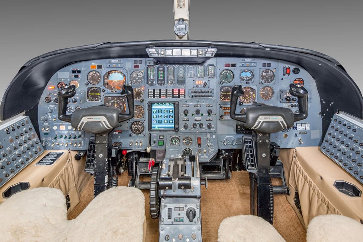 The cockpit of an aircraft with numerous dials, screens, and two control yokes, featuring sheepskin seat covers.