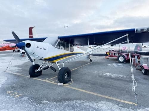 A white Super Cub aircraft with blue and yellow stripes and large tundra tires, parked on a snowy tarmac next to a fuel trailer.