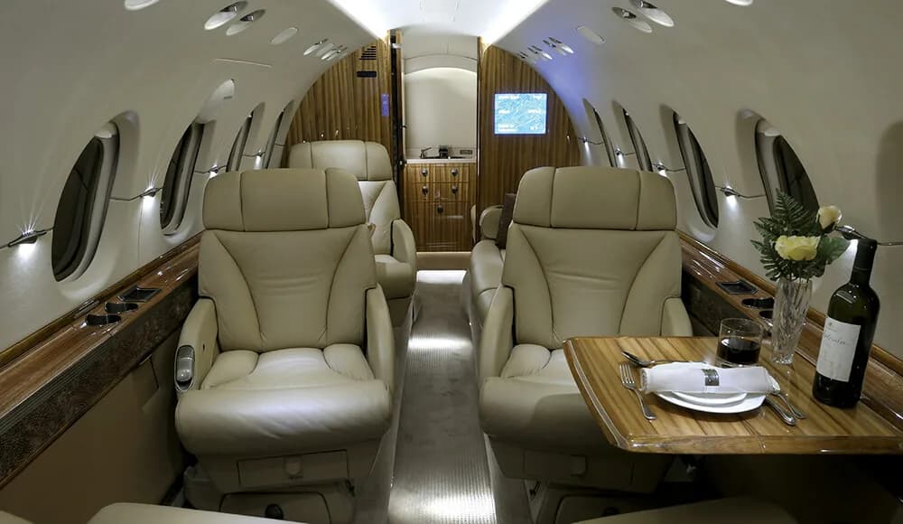 The luxurious interior cabin of a business jet with beige leather seats and a wooden table set for dining.