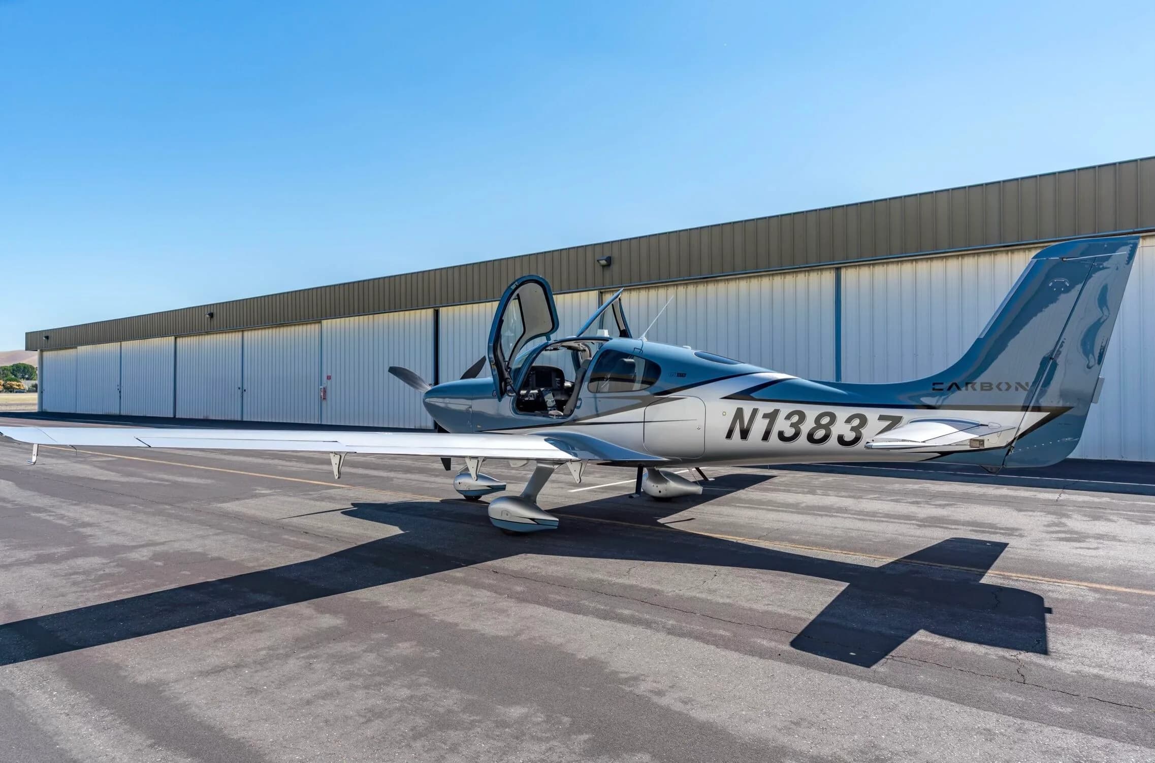 A light blue and silver Cirrus SR22T aircraft, N1383Z, with its doors open, parked on an tarmac.
