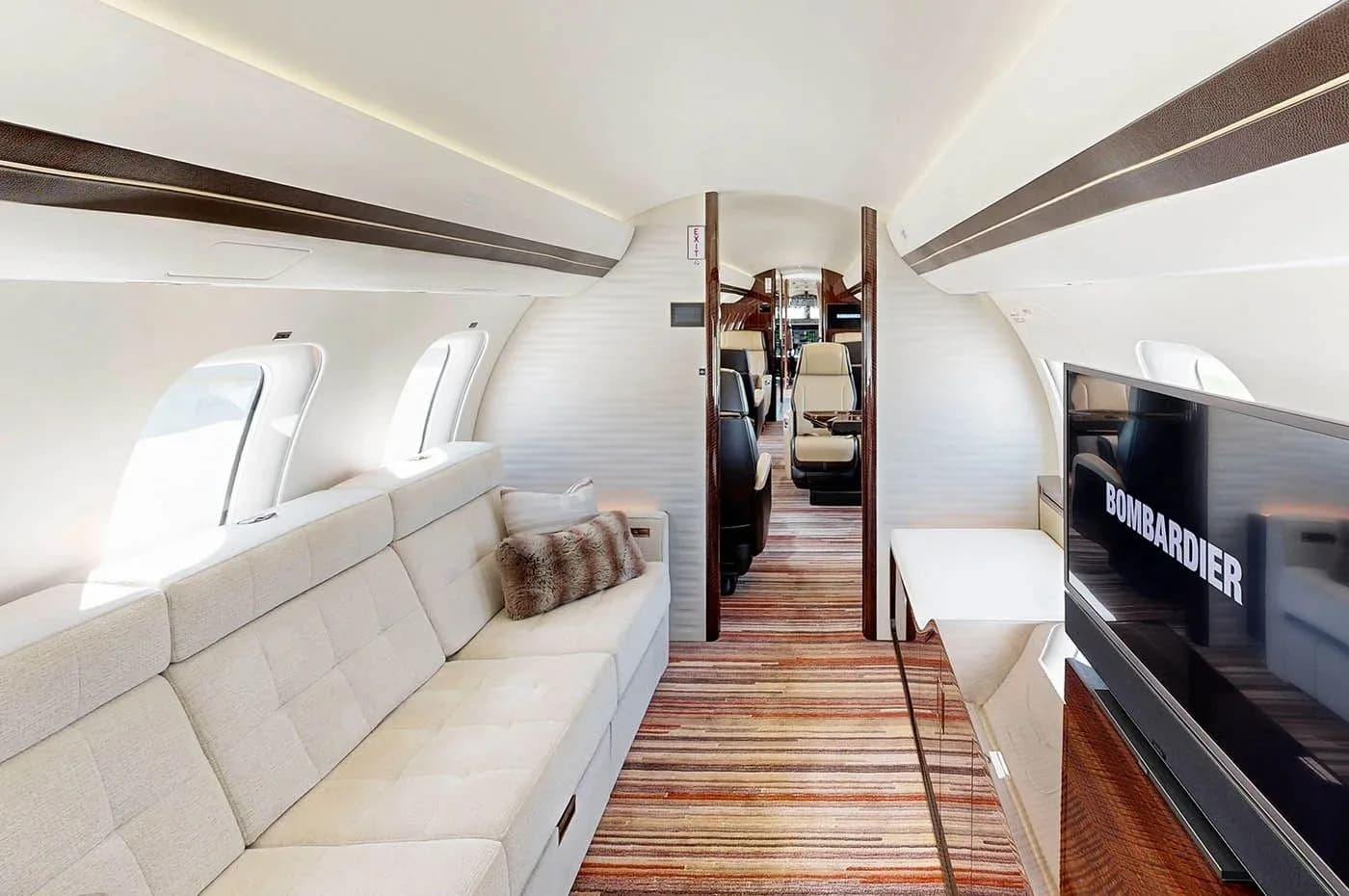 The aft cabin of a Bombardier Global business jet, featuring a long white sofa and a large screen displaying "BOMBARDIER".