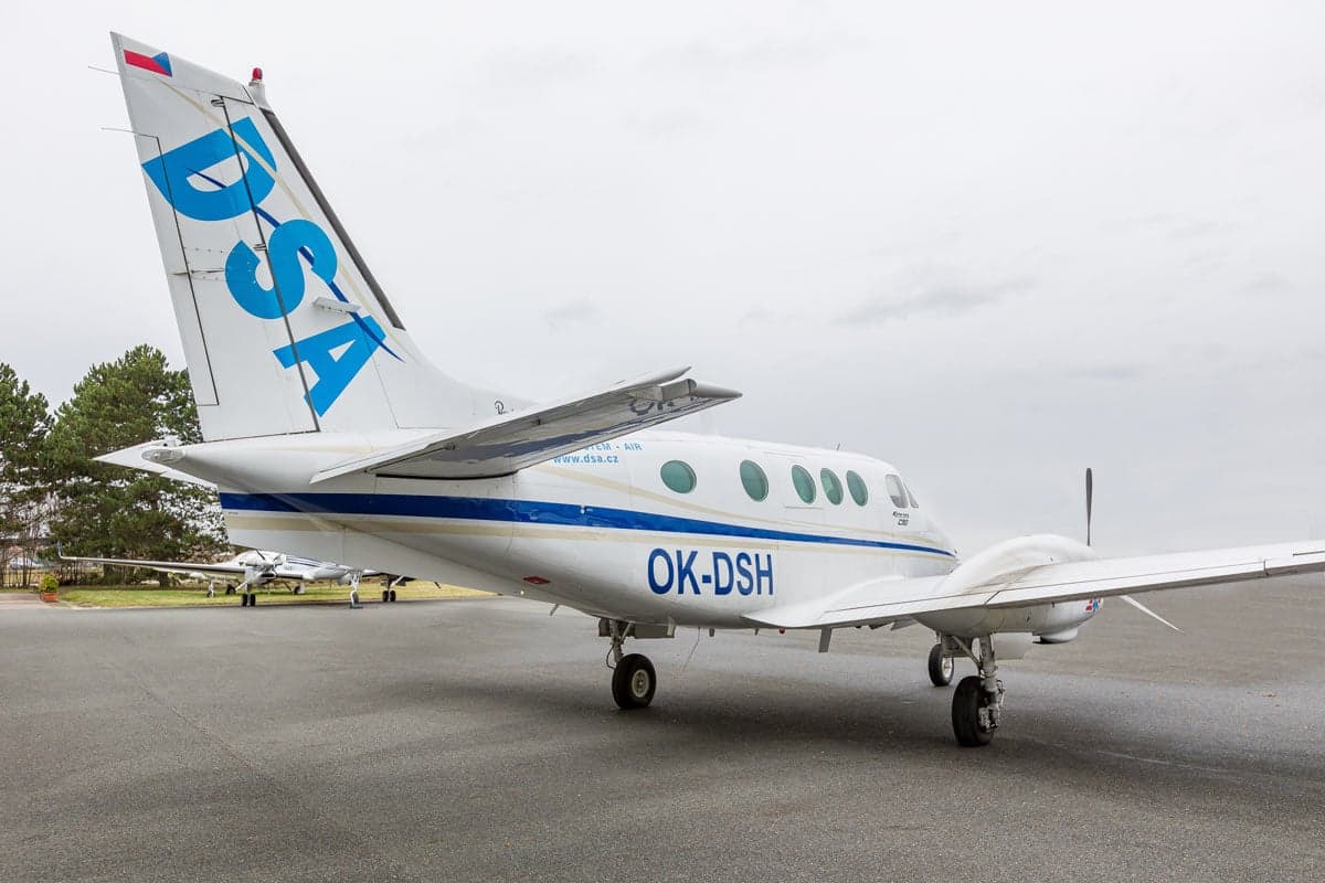 Rear side view of a Beechcraft King Air C90 aircraft with a white and blue paint scheme, parked on the tarmac.