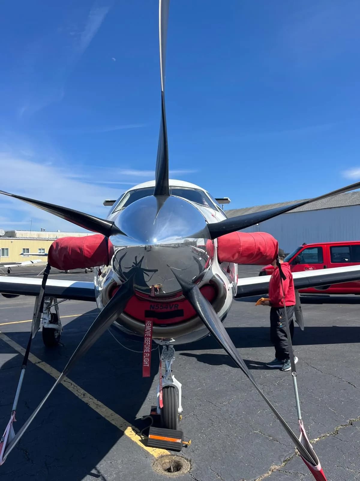 A front view of the Pilatus PC-12 aircraft, showing its propeller with red covers and a person standing nearby.