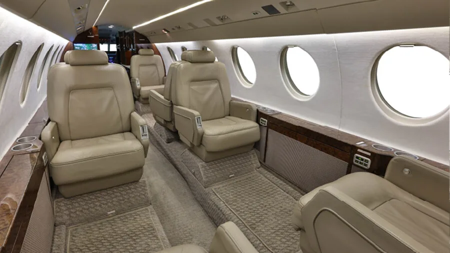 A view down the aisle of a business jet cabin with comfortable beige leather seating and wood accents.