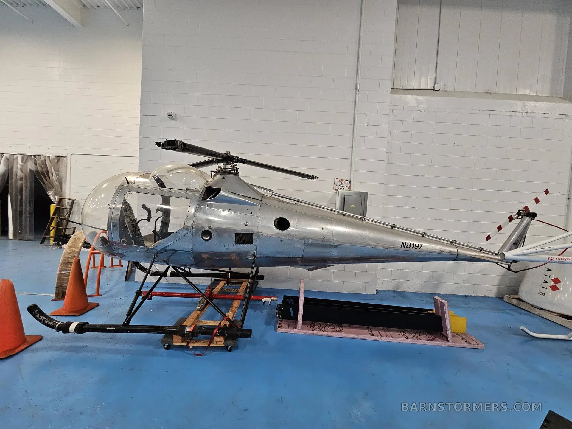 A silver helicopter with registration N8191 is shown from the side, resting on a wooden stand in a hangar.
