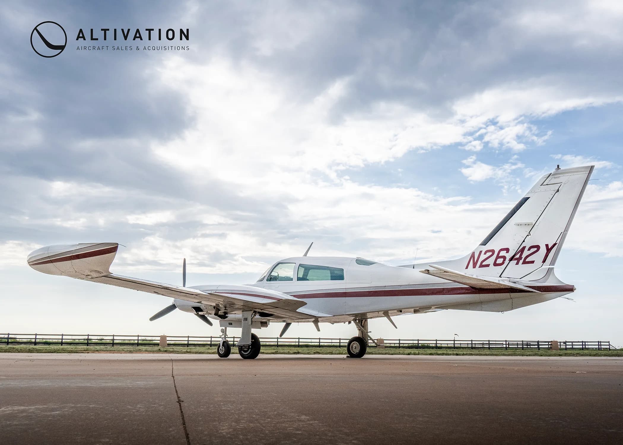 A white twin-engine propeller aircraft with maroon stripes, tail number N2642Y, parked on an airfield under a cloudy sky.