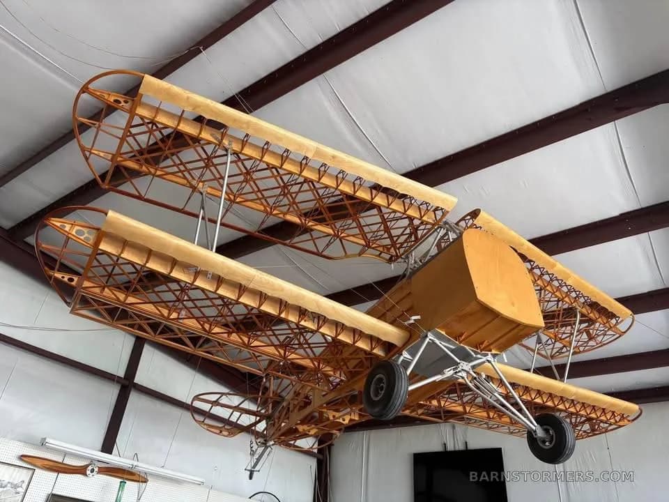 A wooden biplane aircraft frame, with exposed wing and fuselage structure, hangs from the ceiling of a workshop.