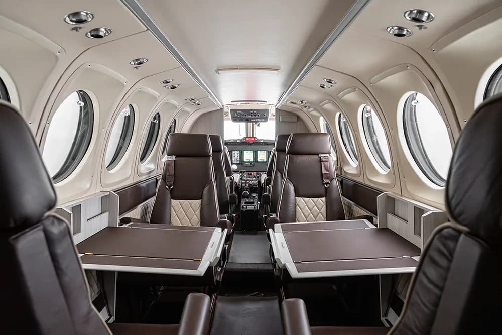 A view down the aircraft cabin aisle, showing brown and beige quilted seats with tables deployed, and the cockpit in the distance.