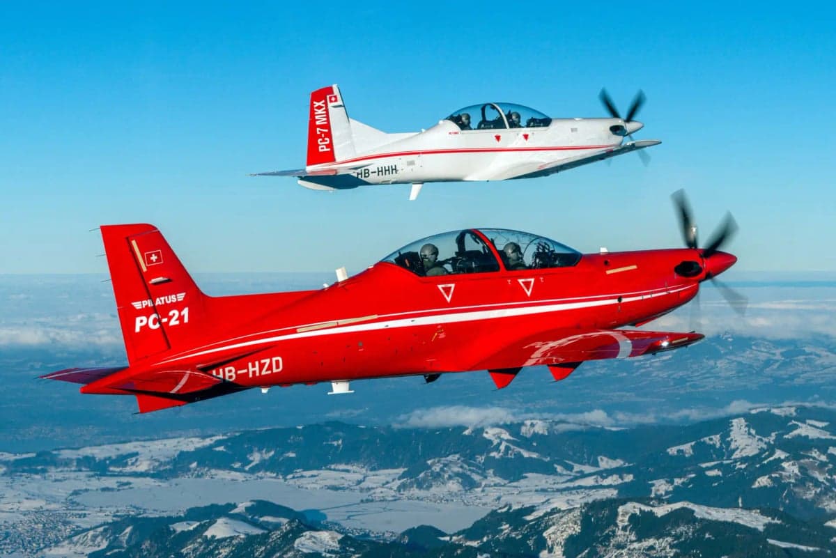 A red Pilatus PC-21 and a white Pilatus PC-7 MKX flying over a snowy mountain range.