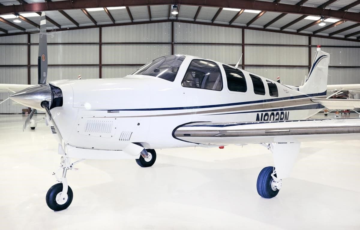 Side-front view of a white Beechcraft Bonanza N802PN with blue and grey stripes, parked in a hangar.