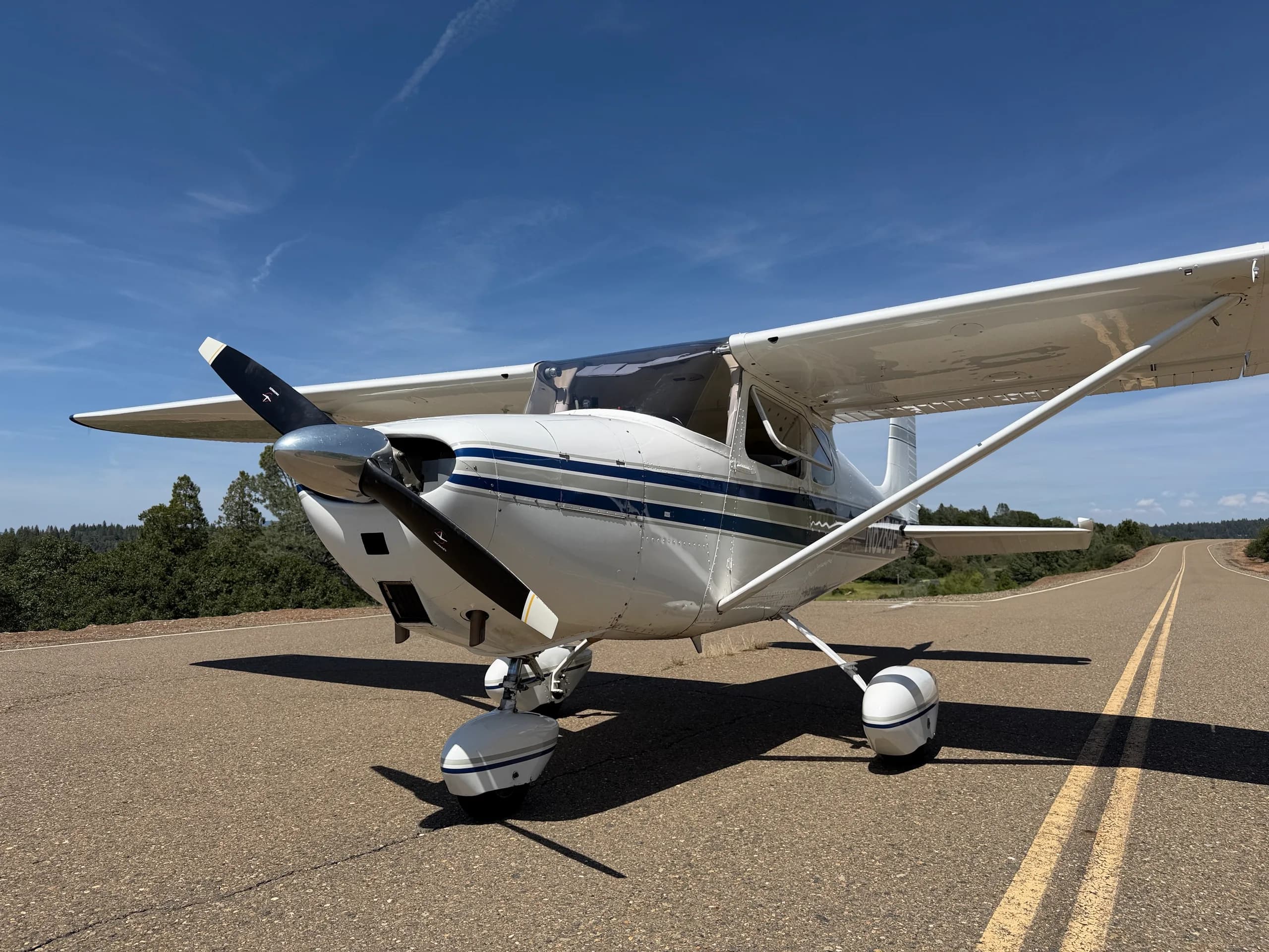 A front-quarter view of a white Cessna 172 with blue stripes, N6284E, showing the propeller and landing gear on an asphalt road.
