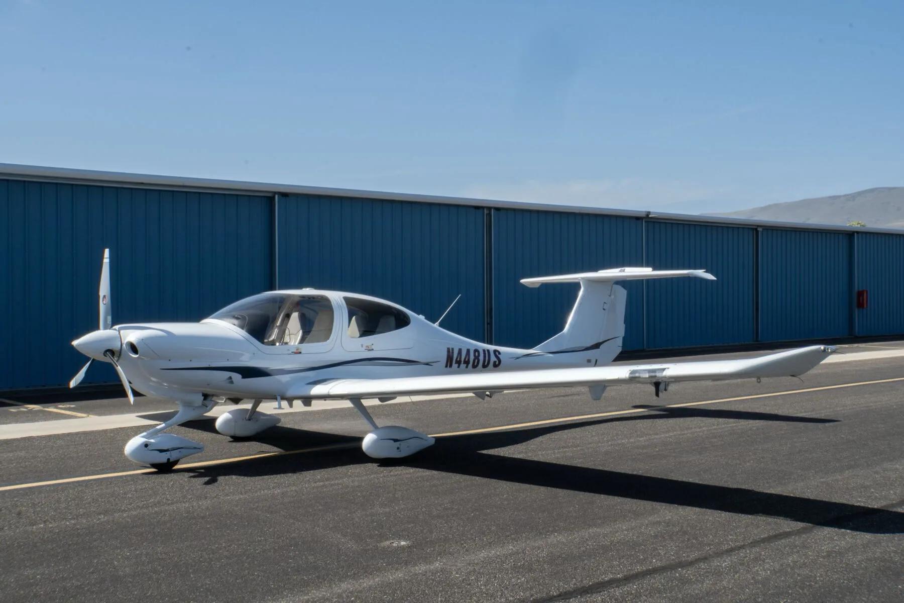 A white Diamond DA40 Star XL aircraft with purple and grey stripes, registration N448US, parked on an tarmac in front of a blue hangar.
