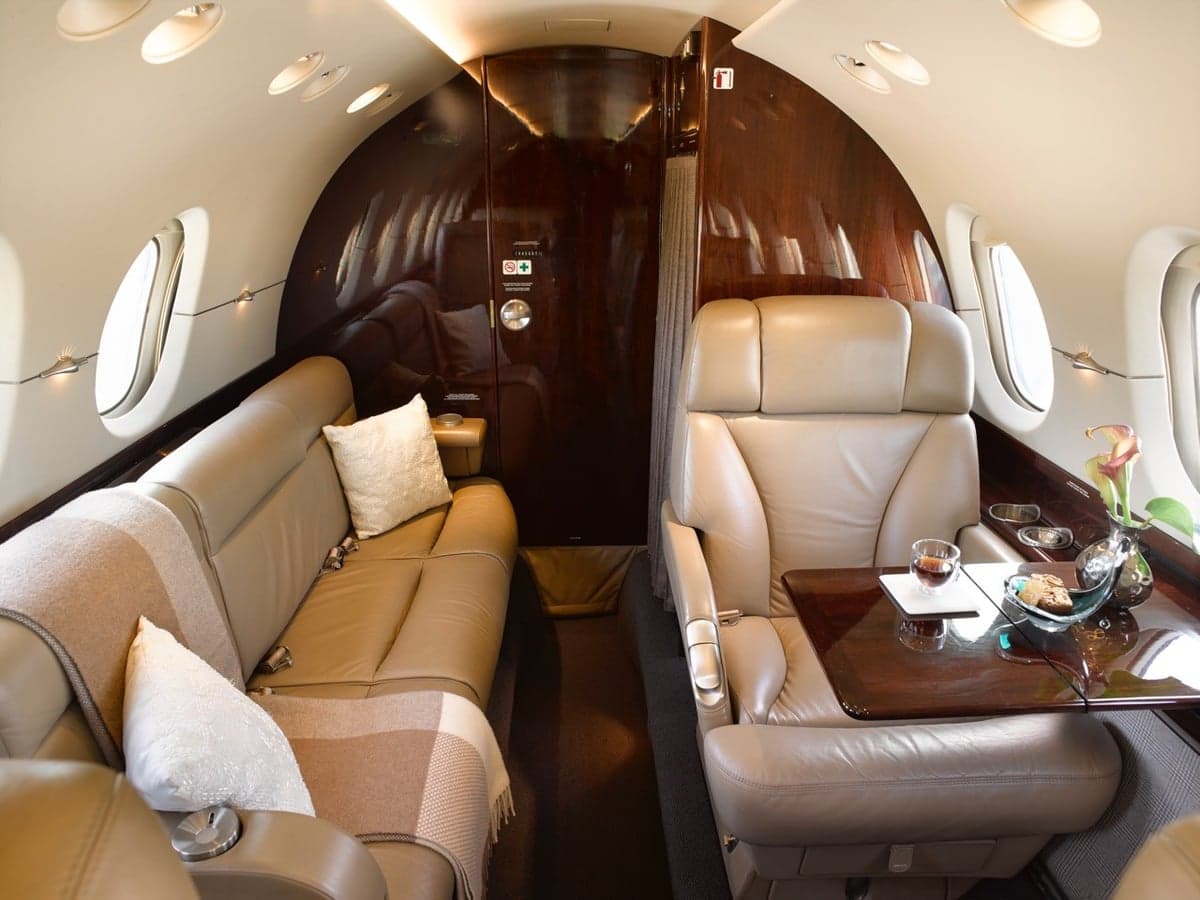 A private jet interior features a tan leather sofa and a single seat with a small table.