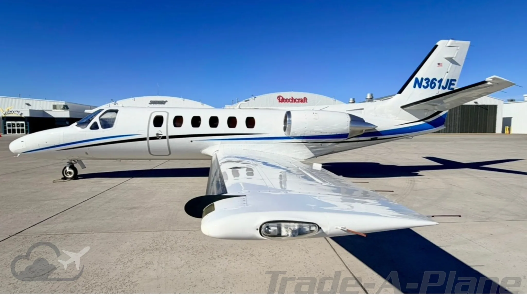 A white business jet with blue stripes and registration N361JE is parked on an airfield, viewed from the front-right side.