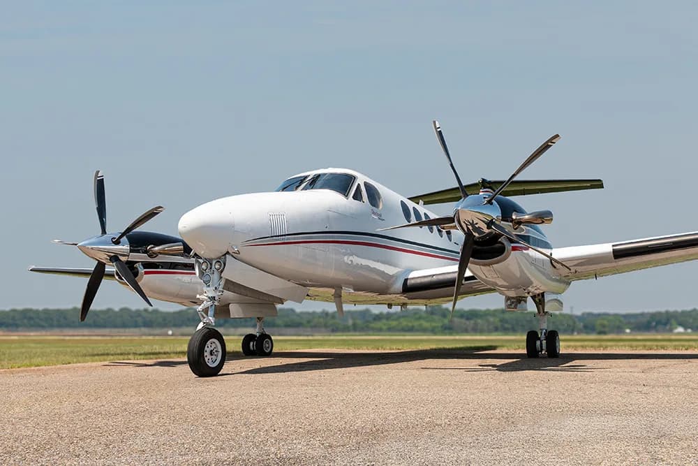 A white twin-turboprop aircraft with black and red stripes parked on an airfield under a clear sky.