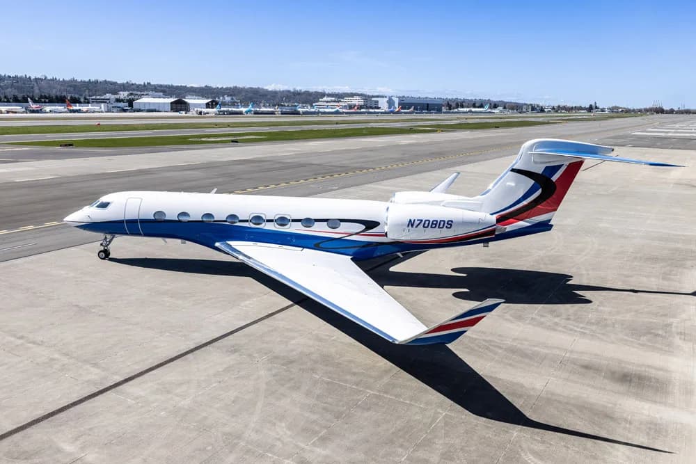 A side profile view of a Gulfstream G650 business jet with a white and blue paint scheme, parked on an airport tarmac.