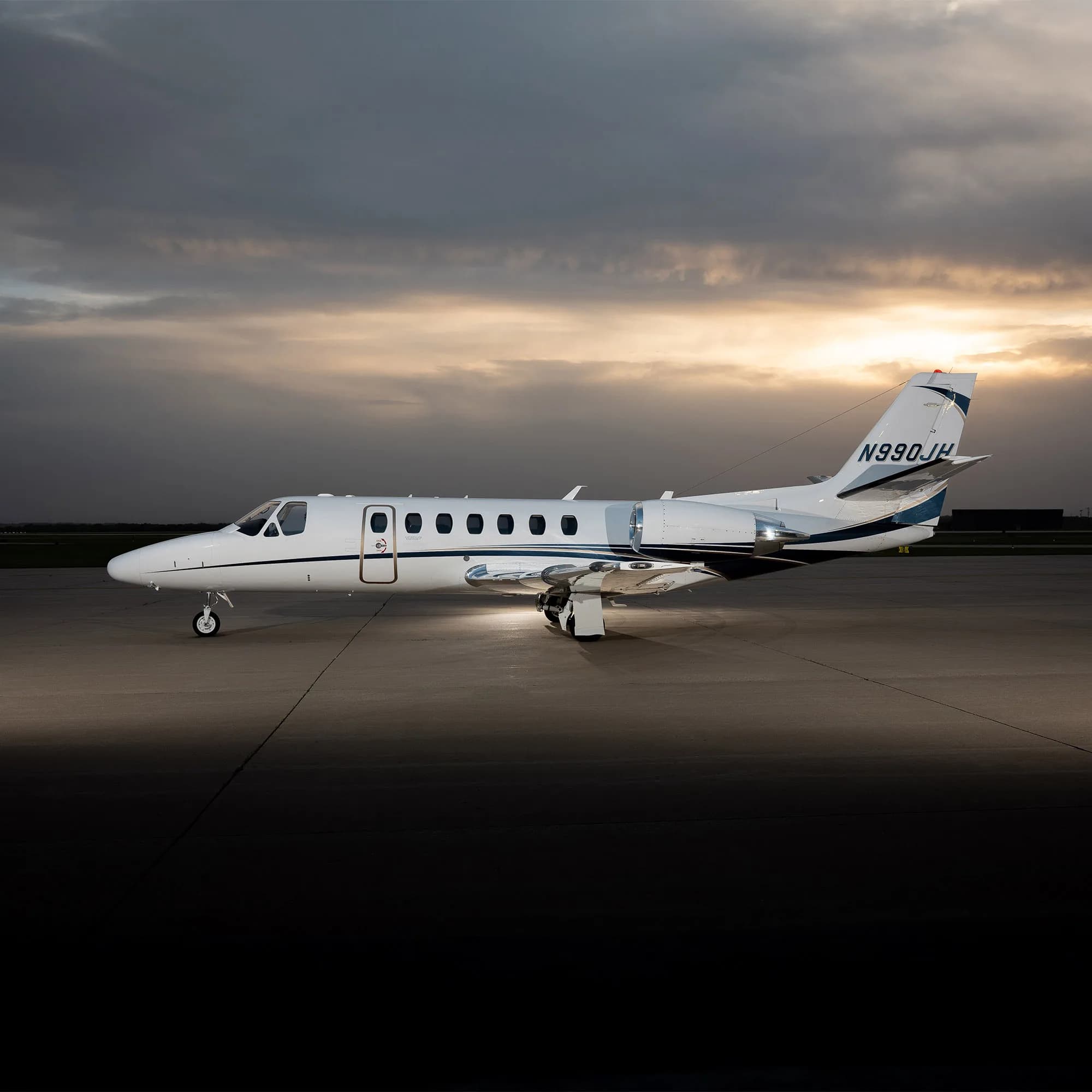 A white Cessna Citation jet with blue and black stripes, registration N990JH, parked on an airfield at sunset.