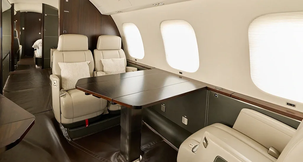 A private jet cabin section featuring cream leather seats and a dark wooden dining table.