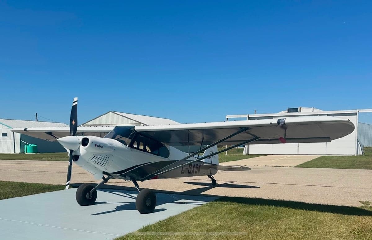 A white, grey, and red CubCrafters XCub aircraft (C-GXSK) parked on an airfield under a clear blue sky.
