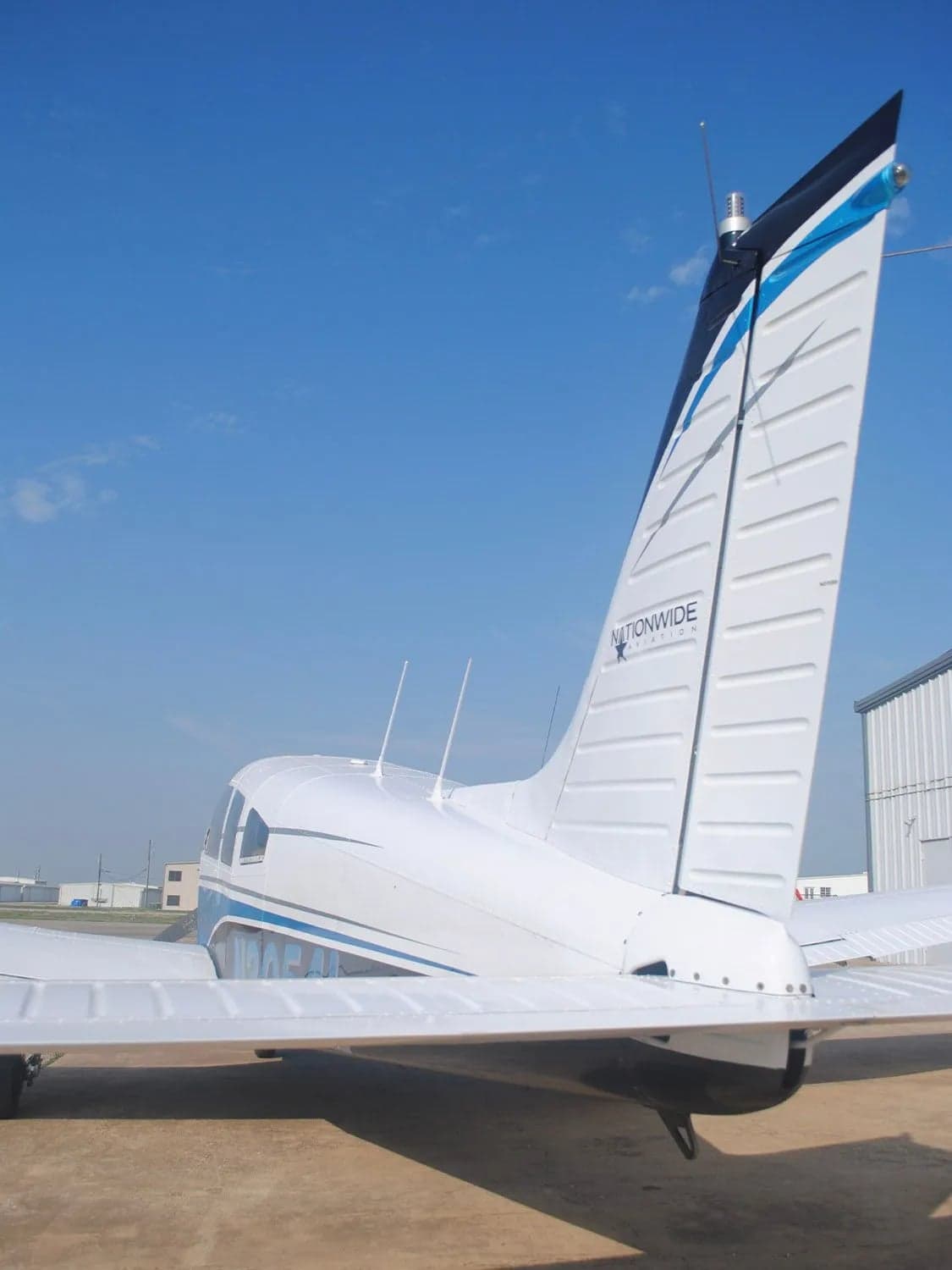 A rear view of a white single-engine aircraft with blue and black tail stripes parked on a tarmac under a clear blue sky.