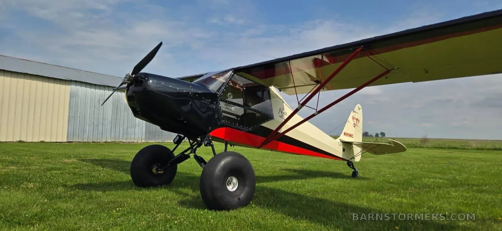 A black, red, and cream-colored bush plane with large tires is parked on a grassy field next to a hangar under a partly cloudy sky.