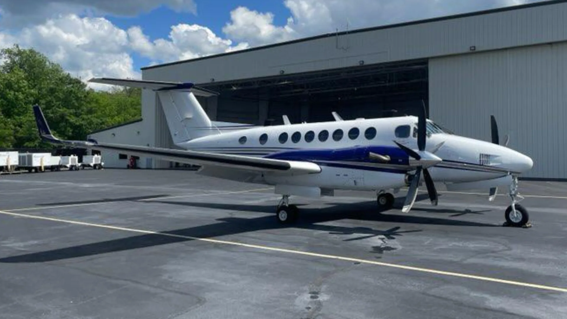 A white and blue Beechcraft King Air N828CJ parked on an asphalt tarmac next to hangars with trees in the background.
