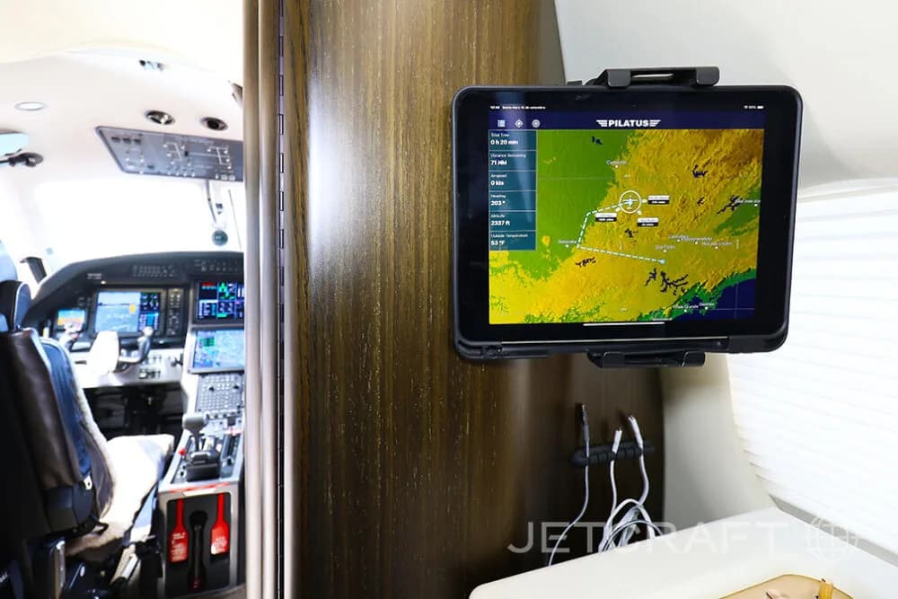 A tablet displaying a flight map mounted on a wooden panel in the cabin of a Pilatus PC-12 aircraft, with the cockpit visible in the background.