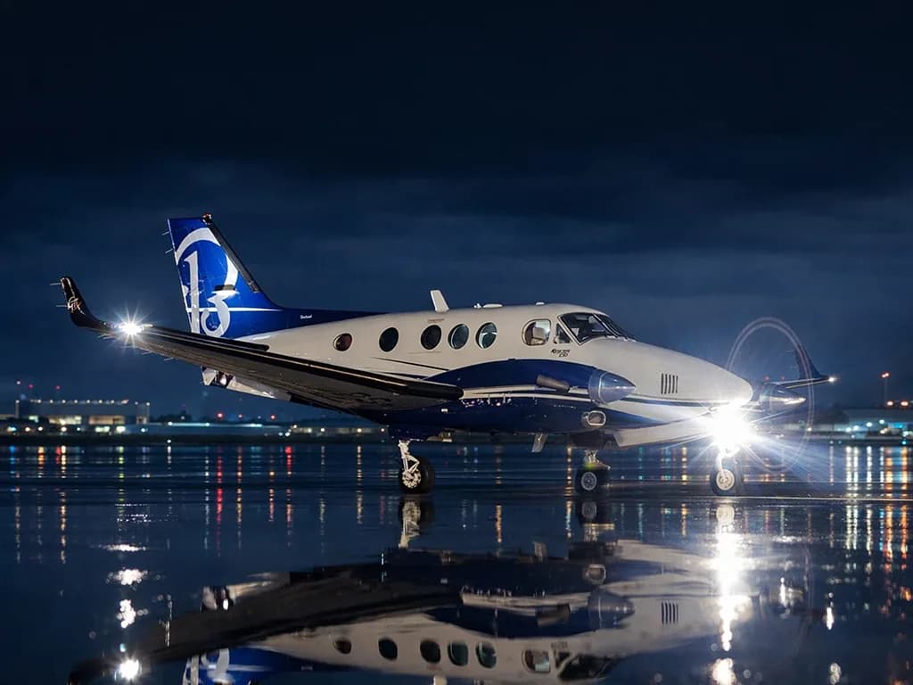 A white and blue twin-turboprop aircraft with a '13' logo on its tail sits on a wet tarmac at night, with landing lights on and city lights reflecting in the water.
