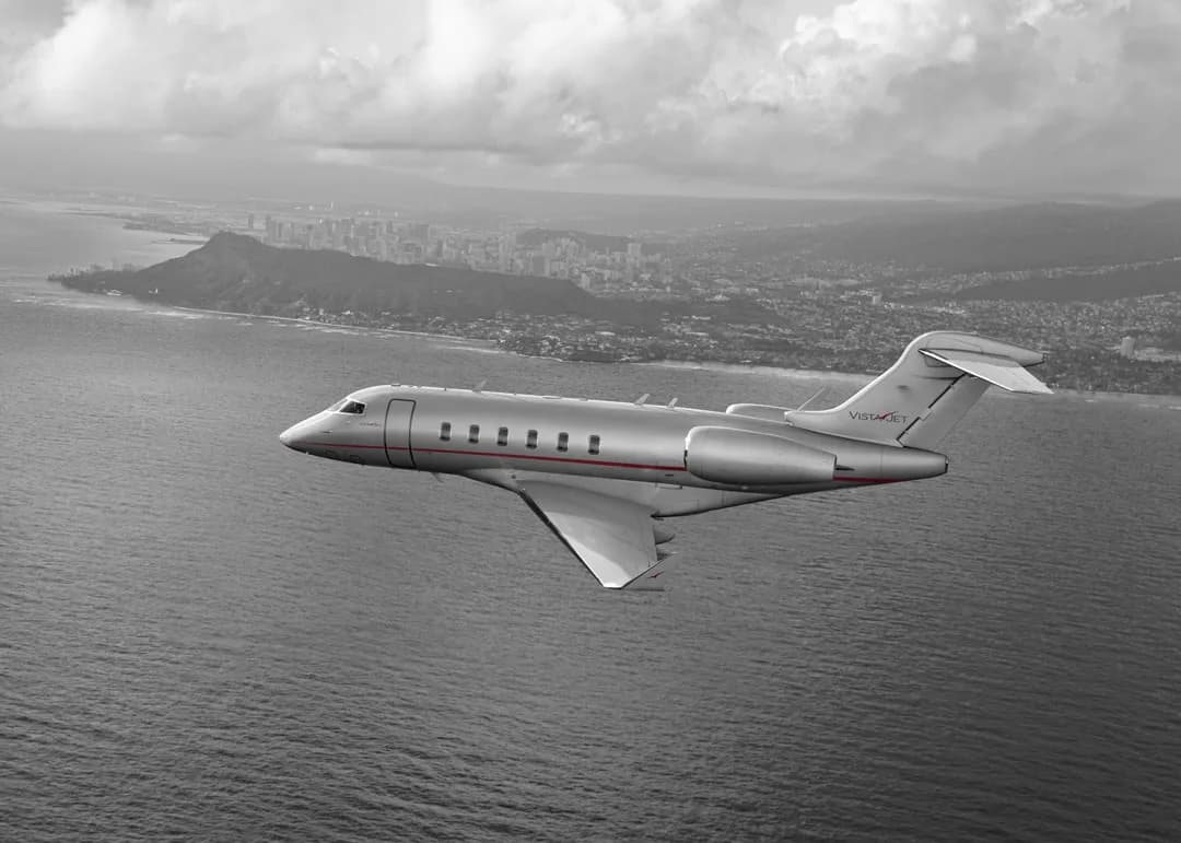 A silver jet flies over the ocean near a city.
