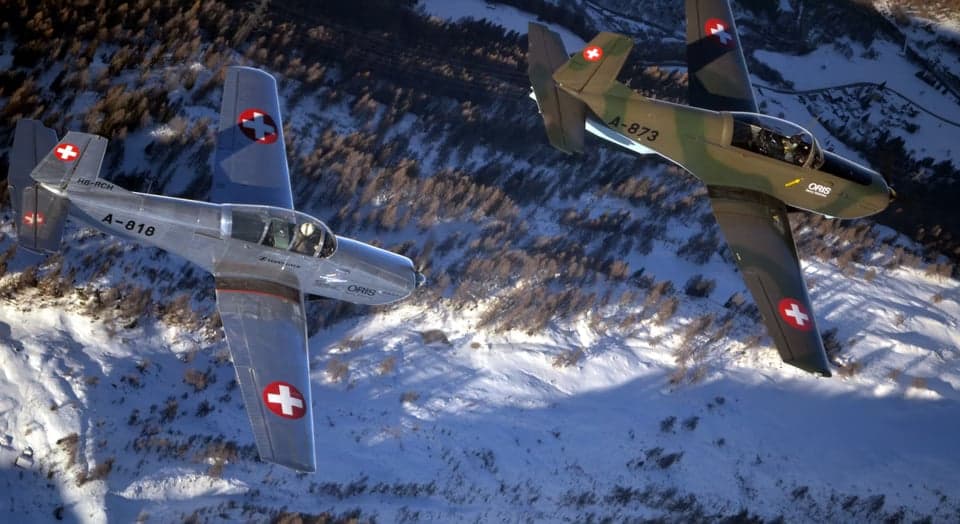 Two Pilatus PC-7 aircraft in flight, one silver and one camouflage.