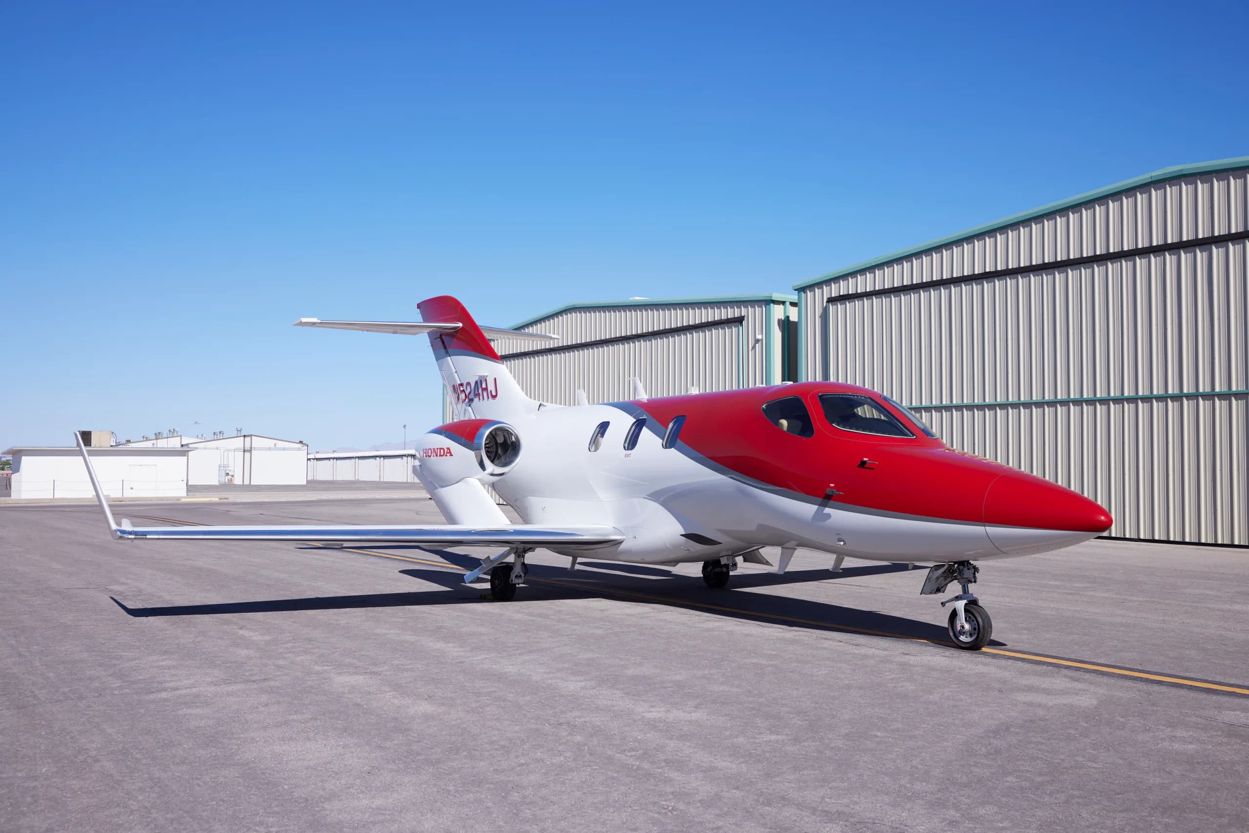 A red and white HondaJet N524HJ parked on an airport tarmac, viewed from the front-right side, with hangars in the background.