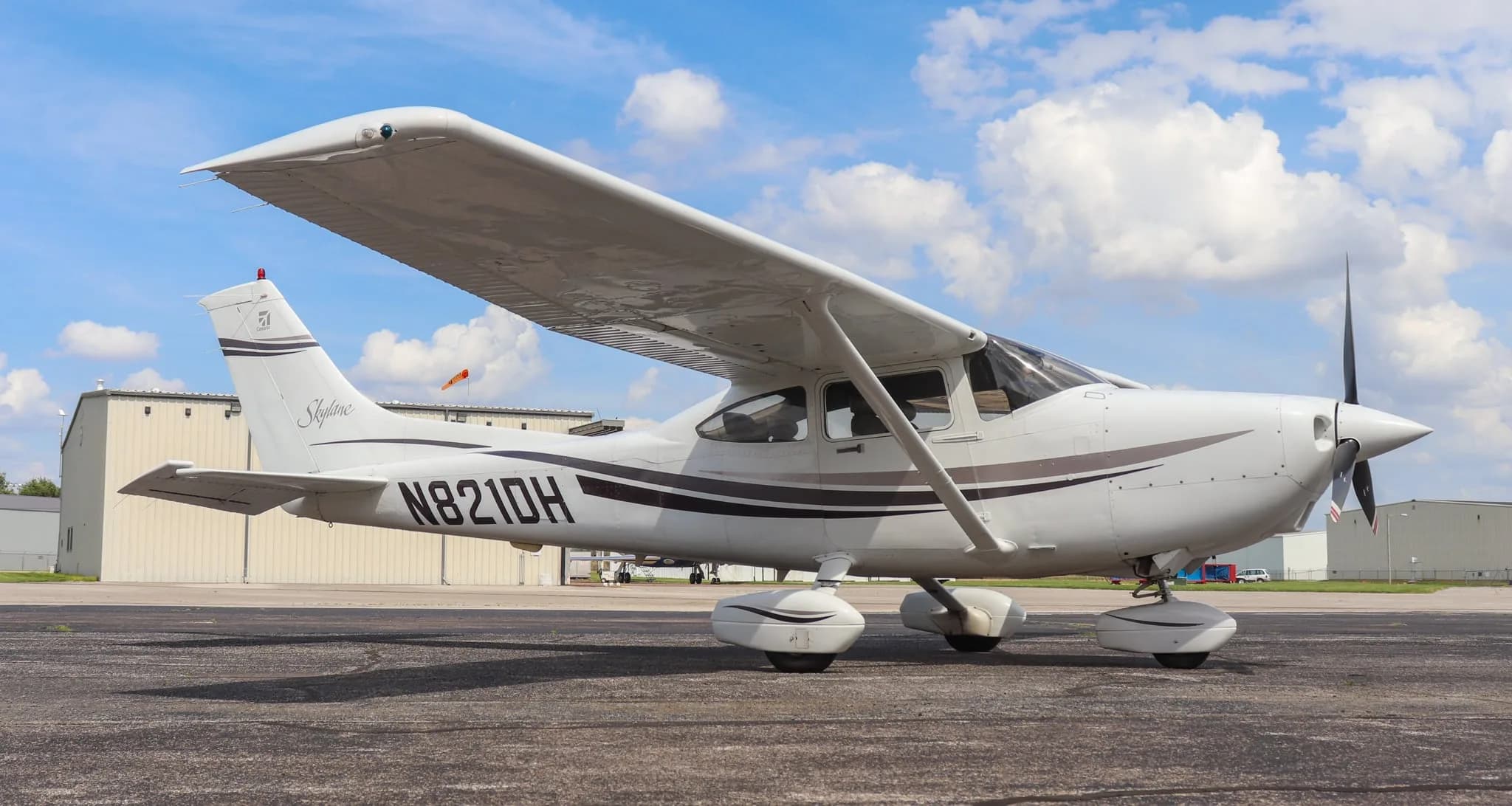 A white Cessna Skylane N821DH with dark grey and brown stripes is parked on an asphalt tarmac under a partly cloudy sky.