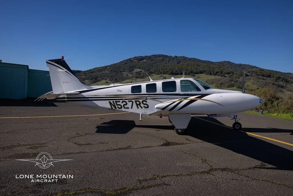 A white Beechcraft Baron N527RS with black and gold stripes parked on an asphalt next to a green hangar and hills.