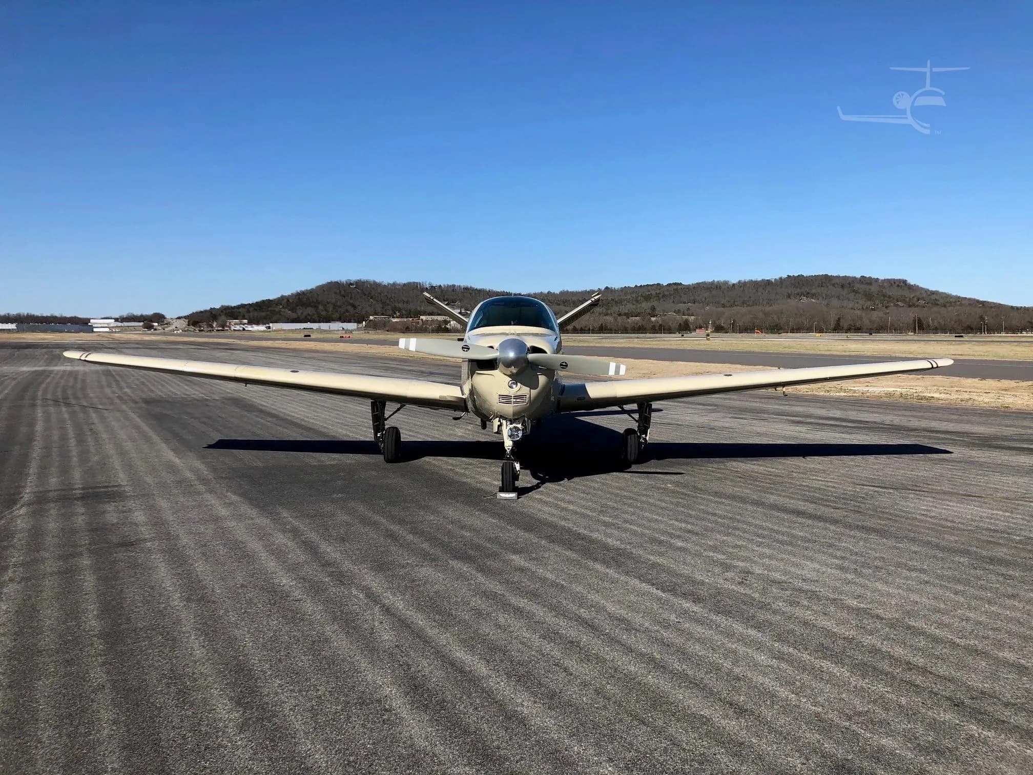 A head-on view of a beige Beechcraft Bonanza N980Q parked on an asphalt runway with hills in the background.