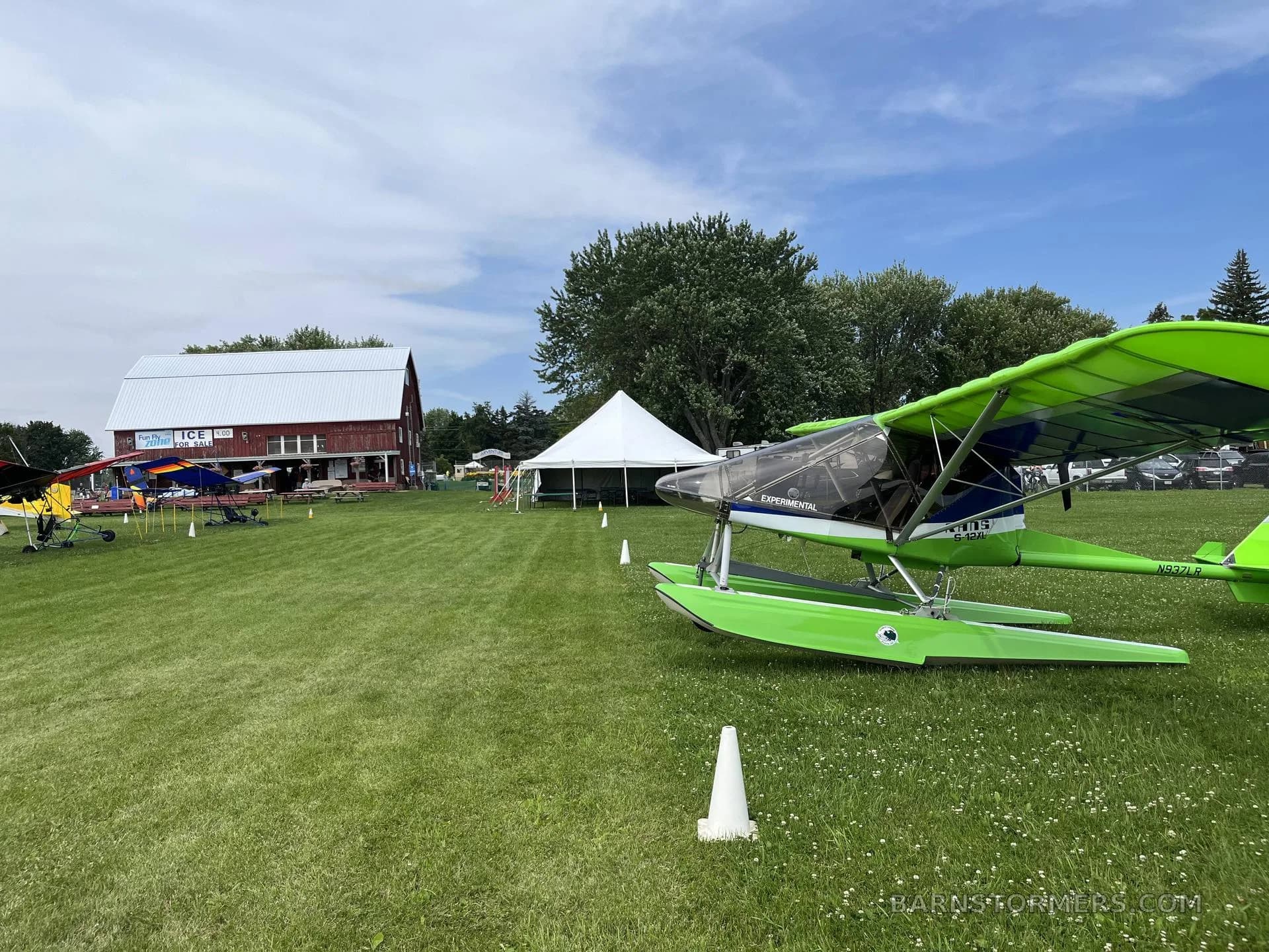 A bright green and blue ultralight seaplane with floats is parked on a grassy field next to a red barn and white tent under a blue sky.