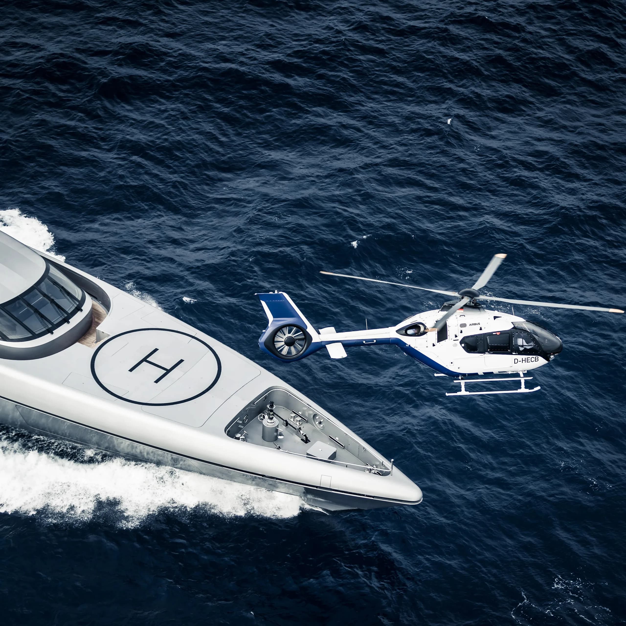 A white and blue Airbus EC135 helicopter flying near a yacht with a helipad.