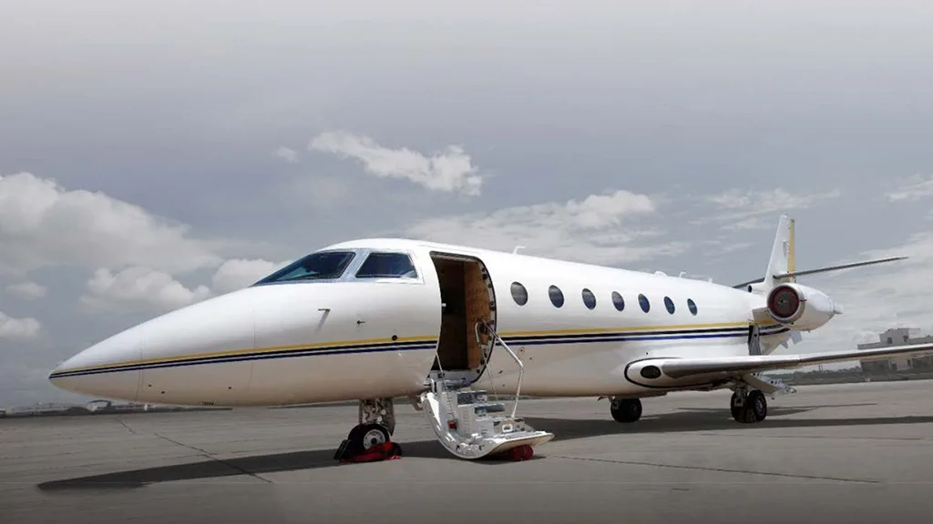 A white private jet with blue and gold stripes on the fuselage, parked on an asphalt tarmac with its main door open and stairs extended.