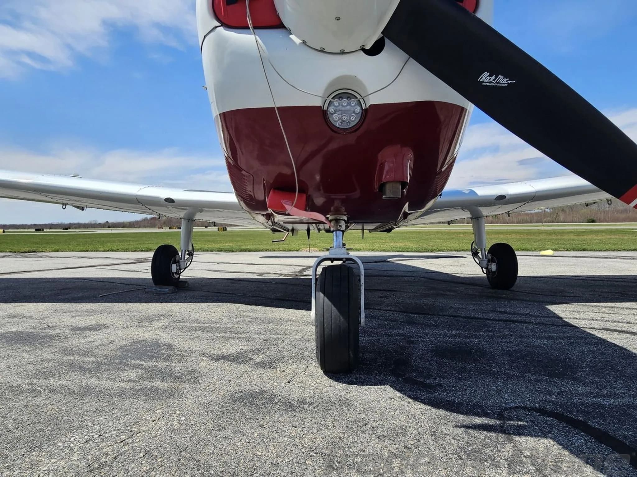 Low-angle view of the front landing gear and propeller of a white and maroon Piper PA-28 aircraft.